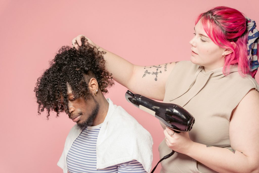 A hairstylist blow-dries a man's curly afro hair in a studio setting.